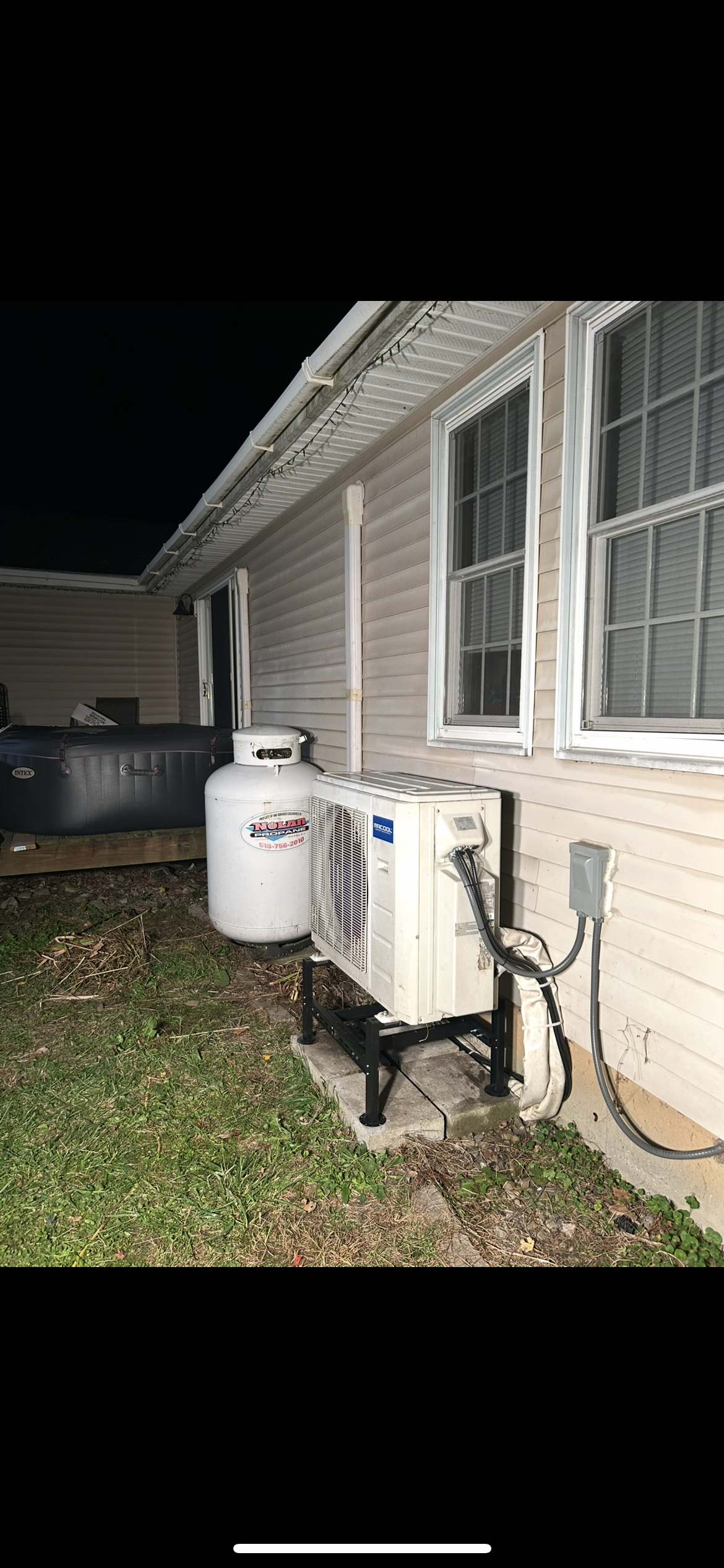 A white outdoor HVAC unit and a propane tank sit on the ground next to the beige vinyl-sided wall of a house at night.