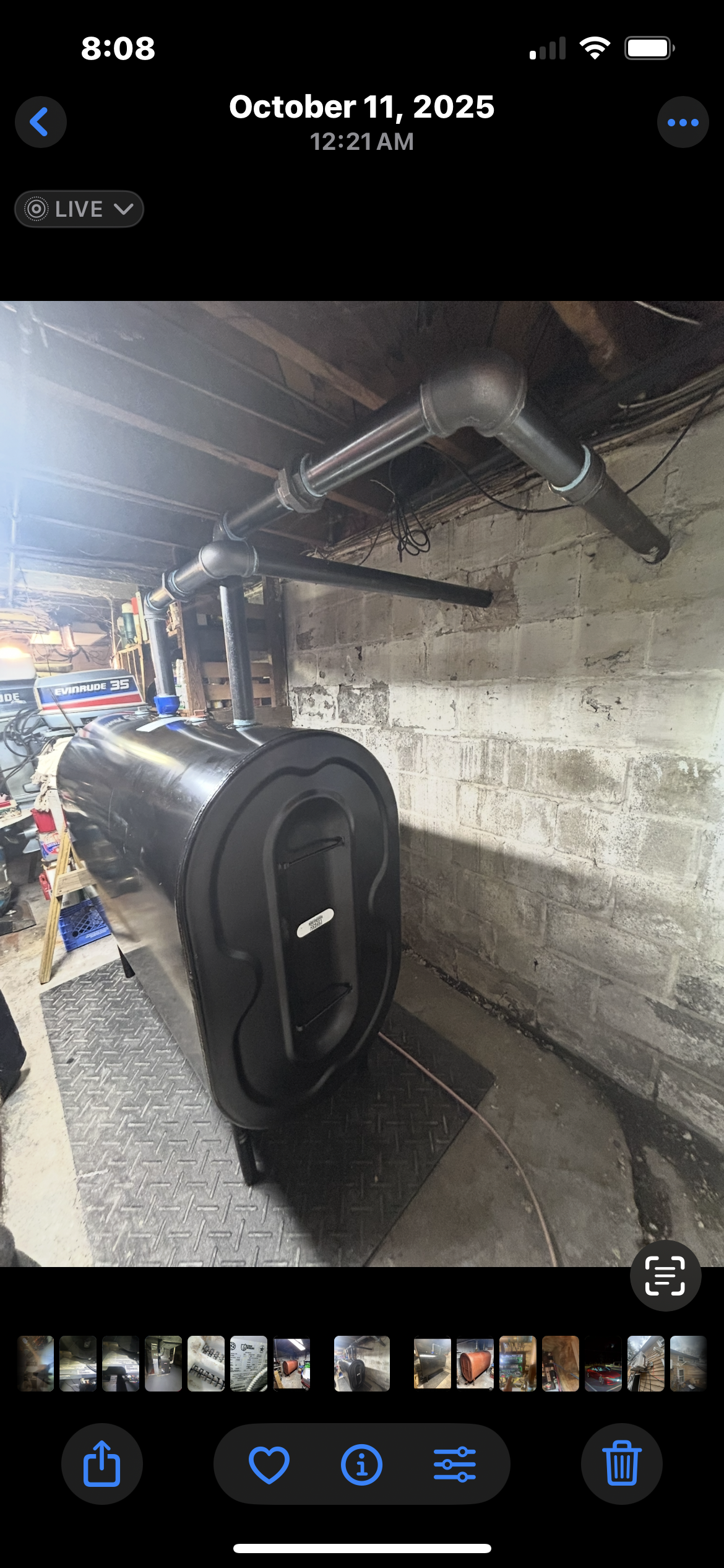 A black furnace sits on a basement floor, with black metal piping running along the unfinished concrete wall above it.