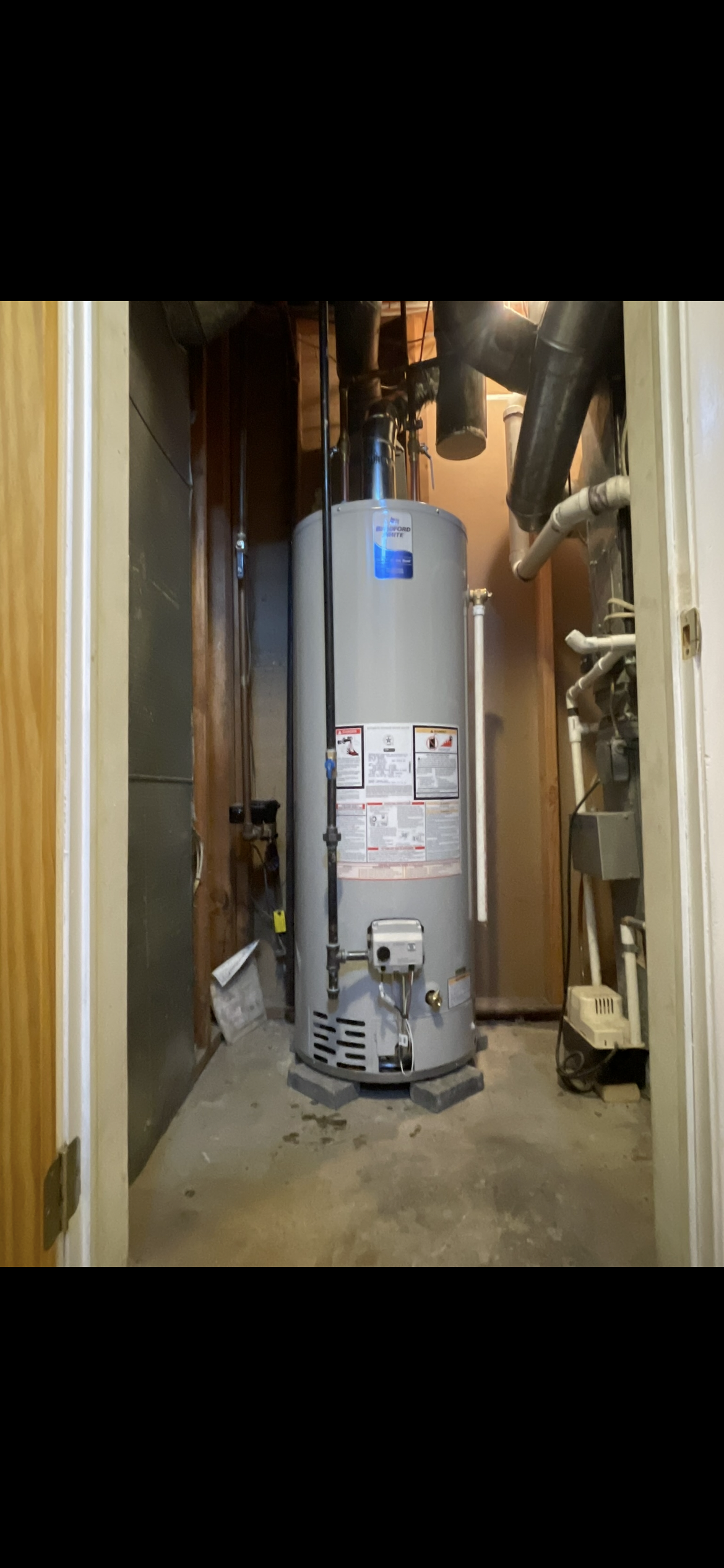 A gray gas water heater stands inside a utility closet, with pipes, valves, and ventilation ducts visible behind it.