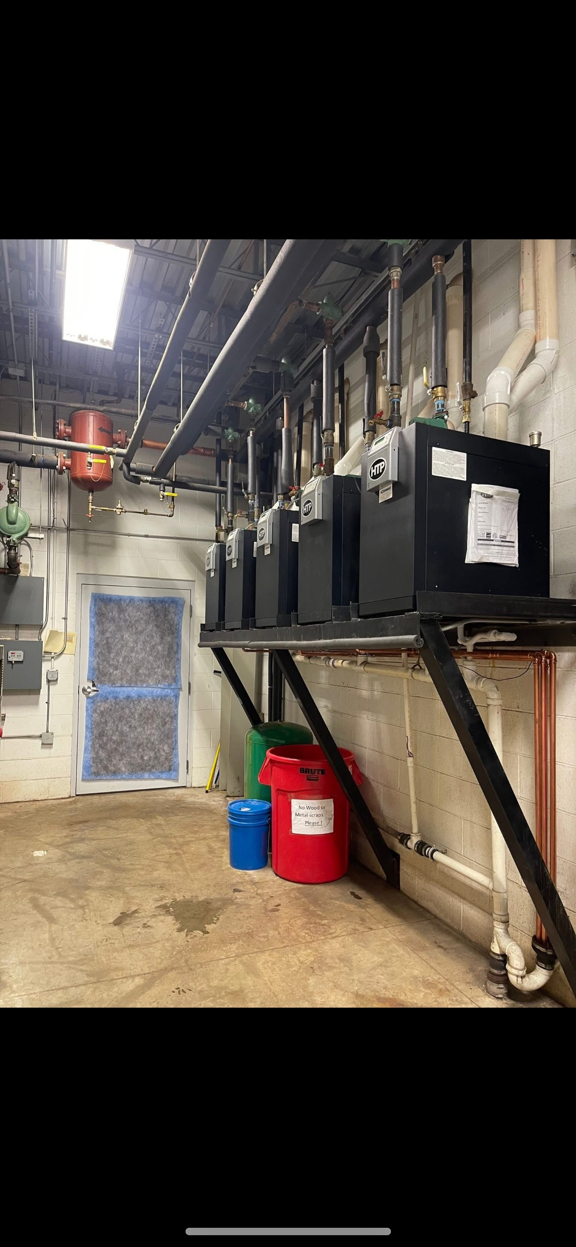 A mechanical room features a wall-mounted row of black utility equipment above red and blue storage bins on the floor.