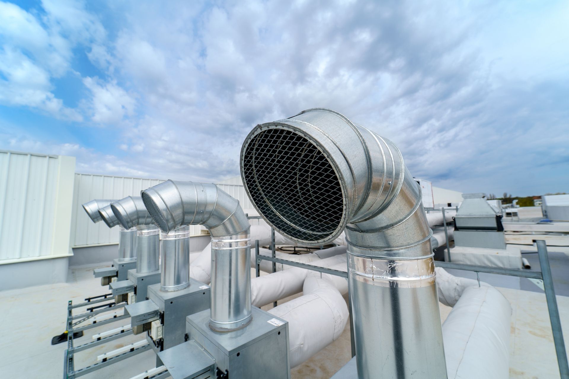 Several metal HVAC exhaust pipes with rounded vents stand on a flat commercial rooftop under a cloudy blue sky.