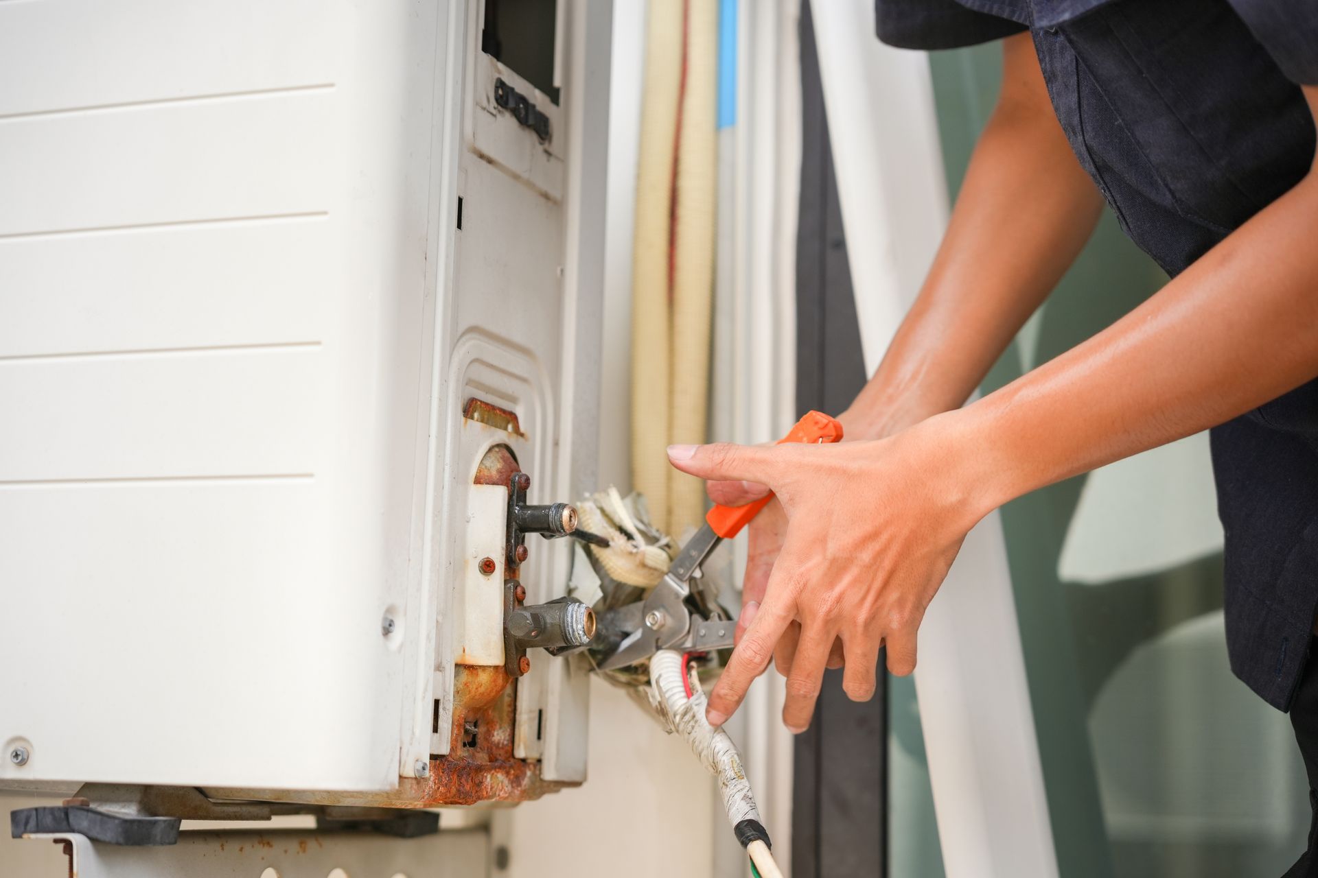 A technician uses pliers to work on the external copper pipes and valves of a white air conditioning unit.