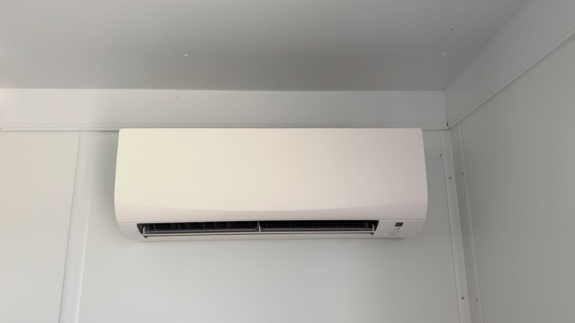 A white wall-mounted air conditioning unit installed high on a white wall in a room.