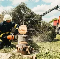 A firefighter in uniform kneels on grass, using a chainsaw to cut a log, with a crane truck in the background.