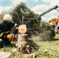 A firefighter in uniform kneels on grass, using a chainsaw to cut a log, with a crane truck in the background.