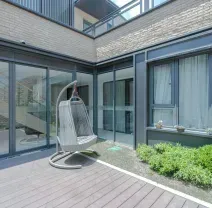 A modern courtyard with wood decking, a hanging egg chair, and glass-paneled walls surrounding a small patch of greenery.