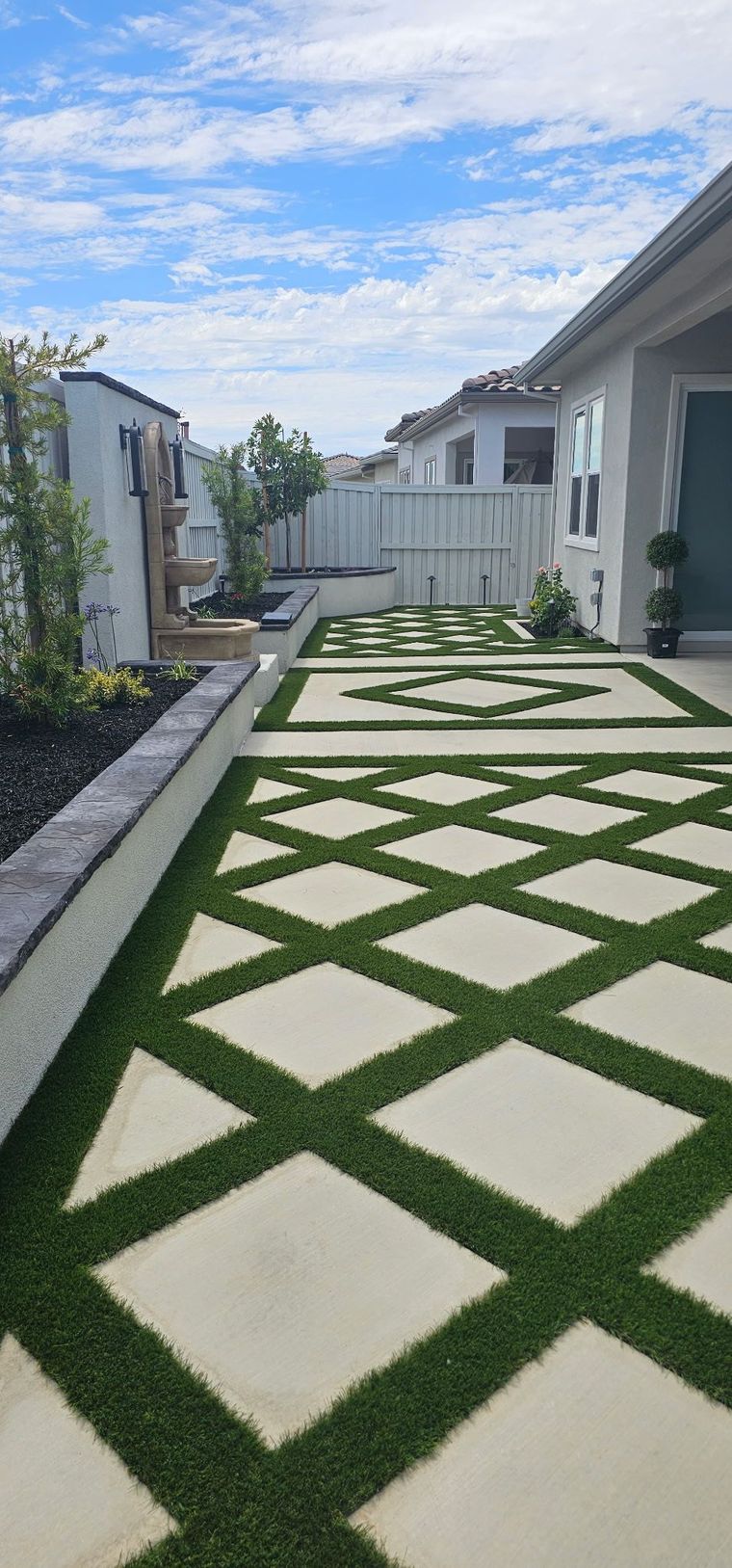 A walkway made of geometric concrete pavers with artificial grass between them, alongside a house exterior and garden.