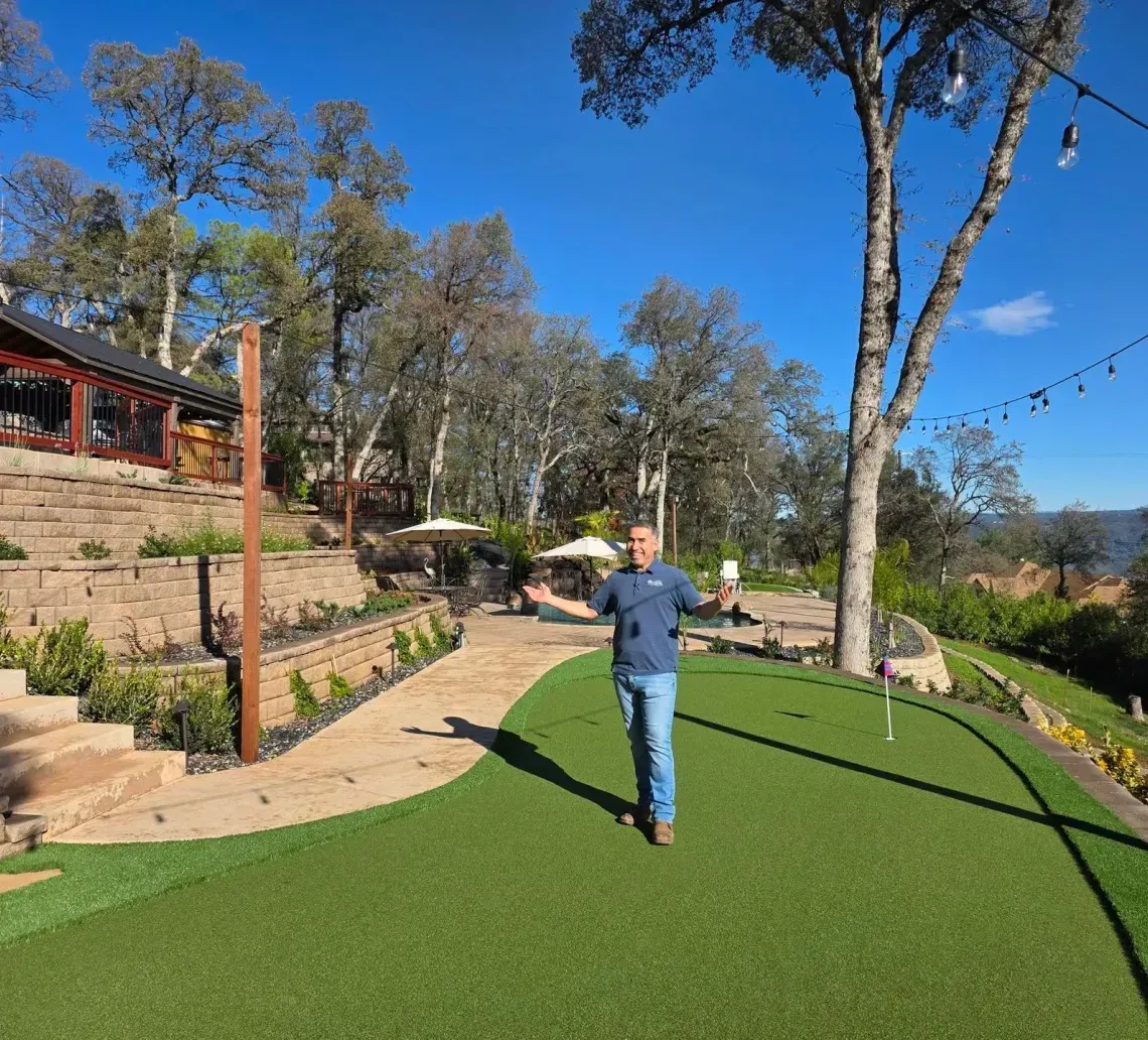 A person stands on a green turf lawn in front of a stone-terraced outdoor patio area with trees under a blue sky.