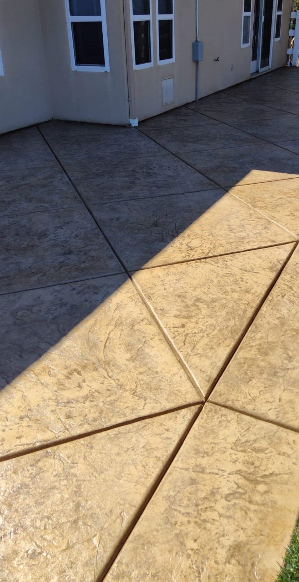 A view of a tan concrete patio with a triangular pattern, partially shaded, beside a tan house with windows.