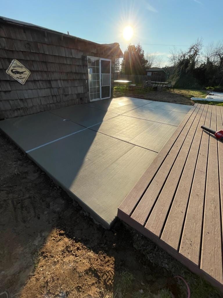 A freshly poured concrete patio adjacent to a wood-plank deck and the side of a brown-shingled building in the sunlight.