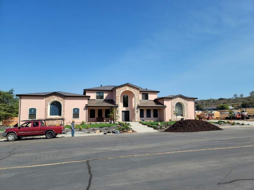 A large, two-story pink house with stone accents stands under a clear blue sky, with a red truck parked in the foreground.