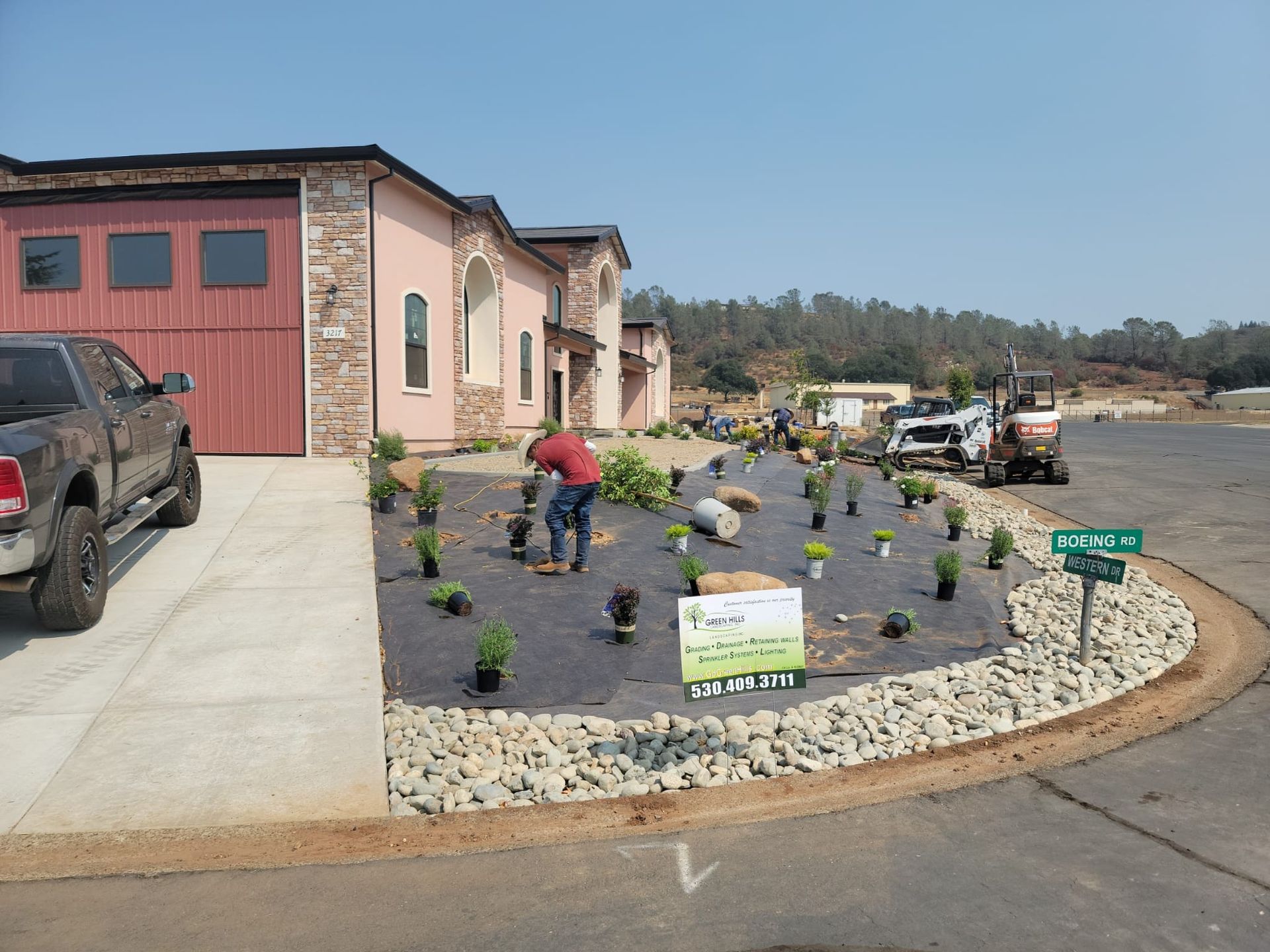 A person plants small greenery in a mulched landscaping bed in front of a house, with a truck parked on the driveway.
