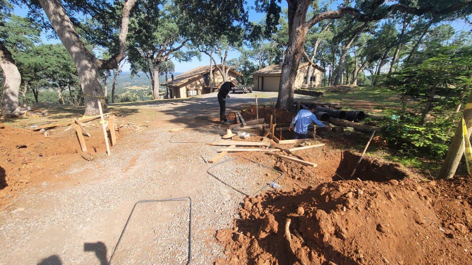 Two people work on an excavation project with wooden forms, soil, and gravel near trees and houses on a sunny day.