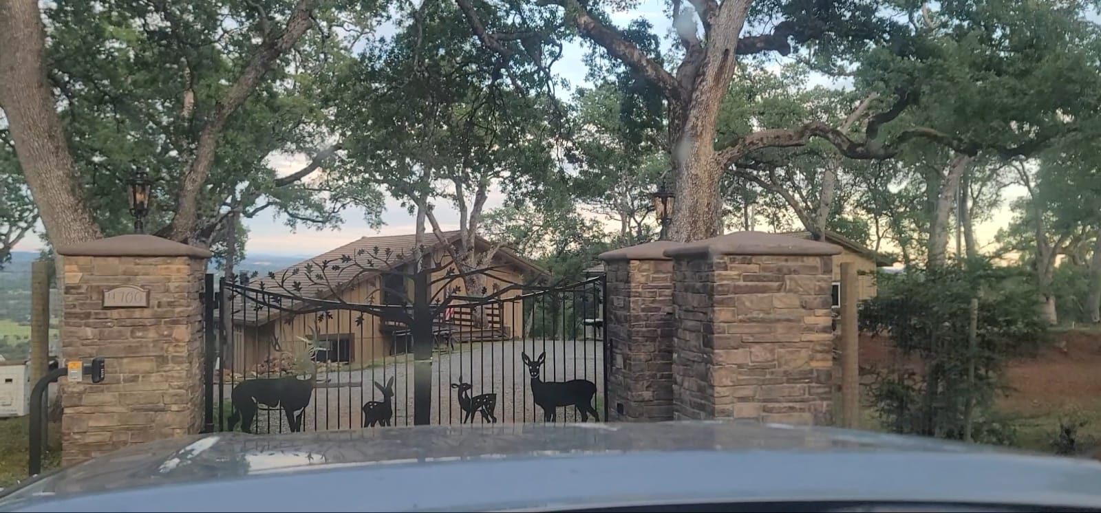 A stone-pillared gate with metal deer silhouettes sits in front of a rustic home surrounded by trees at sunset.