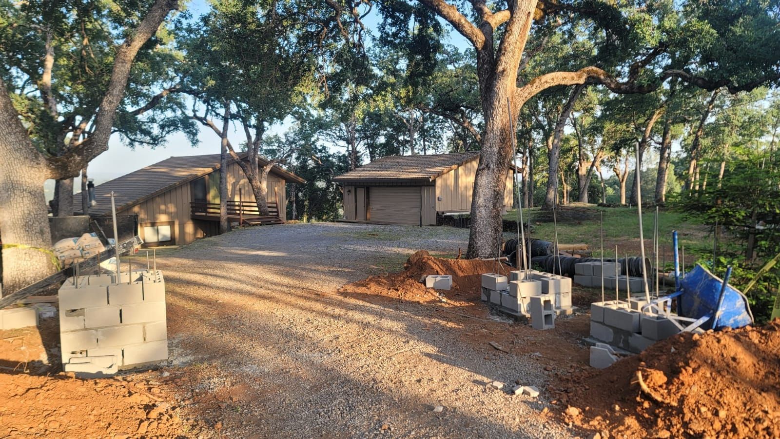 Construction site with partially built concrete block pillars and rebar in a wooded yard with houses in the background.