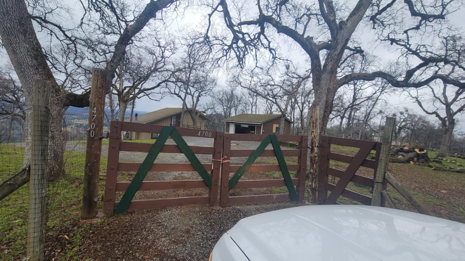 A pair of wooden entrance gates with green cross-bracing stand in a rural setting, with buildings visible in the distance.