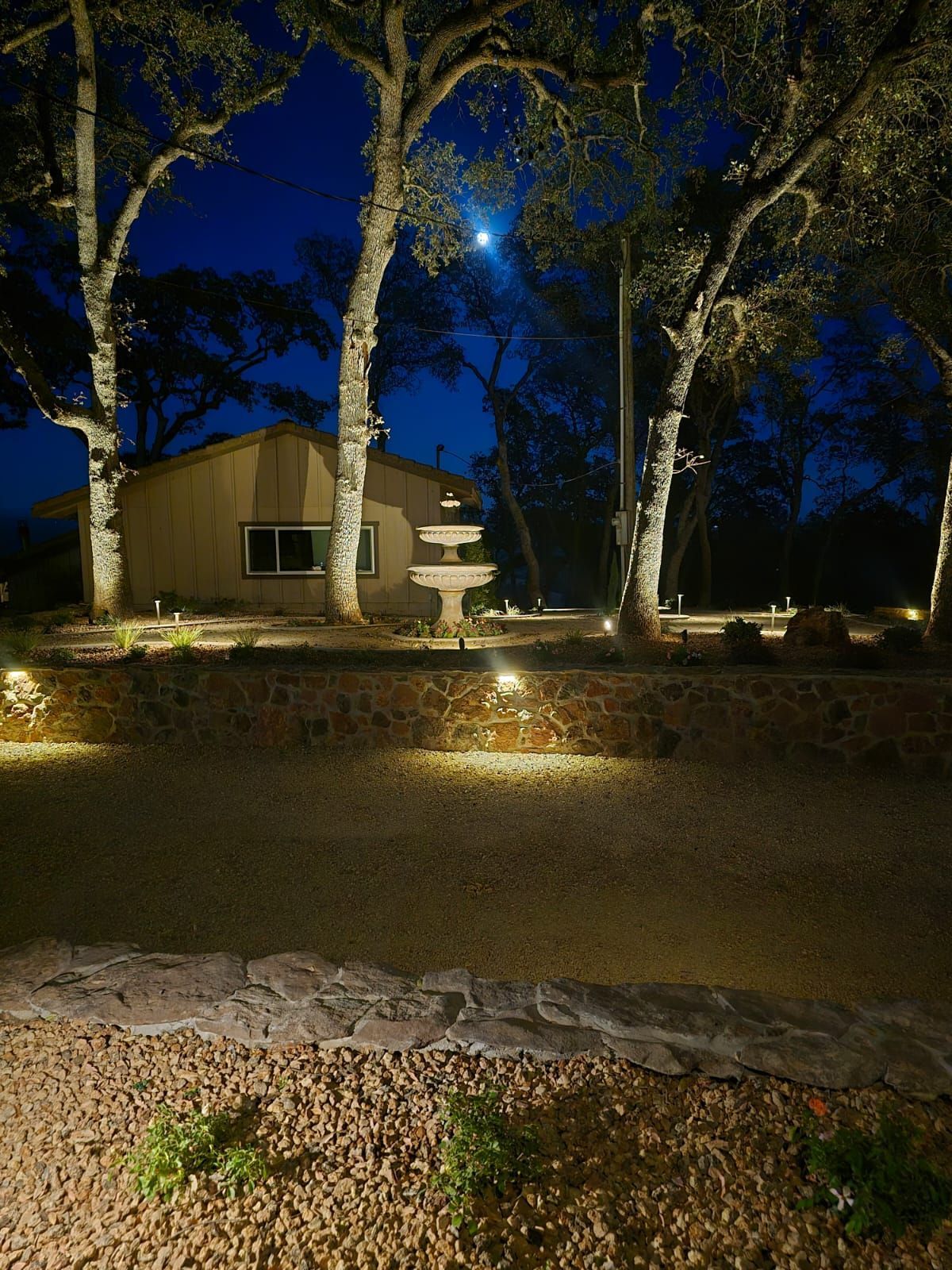 An outdoor home scene at twilight with illuminated landscaping, a tiered fountain, and a gravel yard.