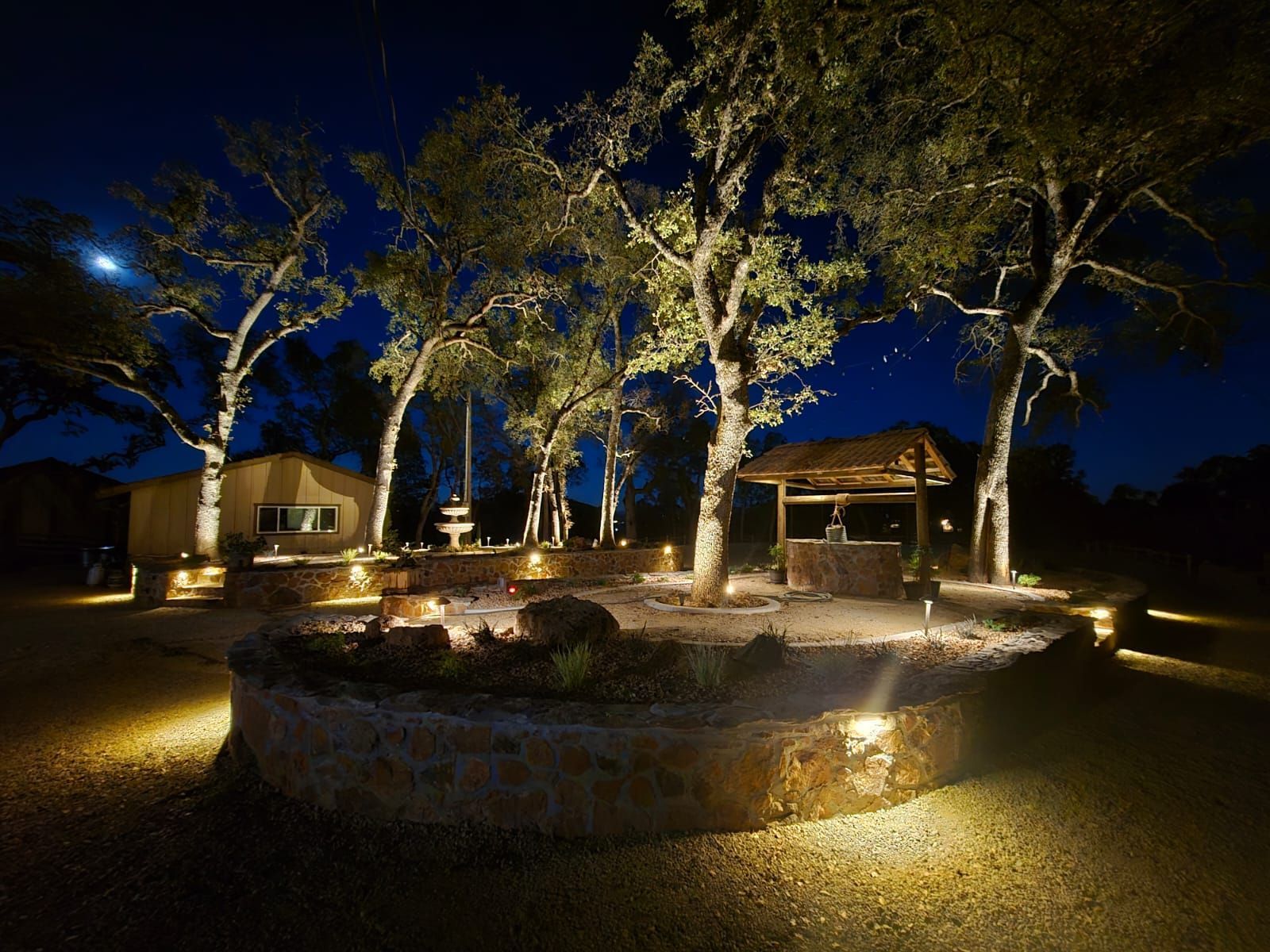 Night scene of a stone-walled courtyard landscaped with illuminated trees, a small structure, and warm ambient lighting.