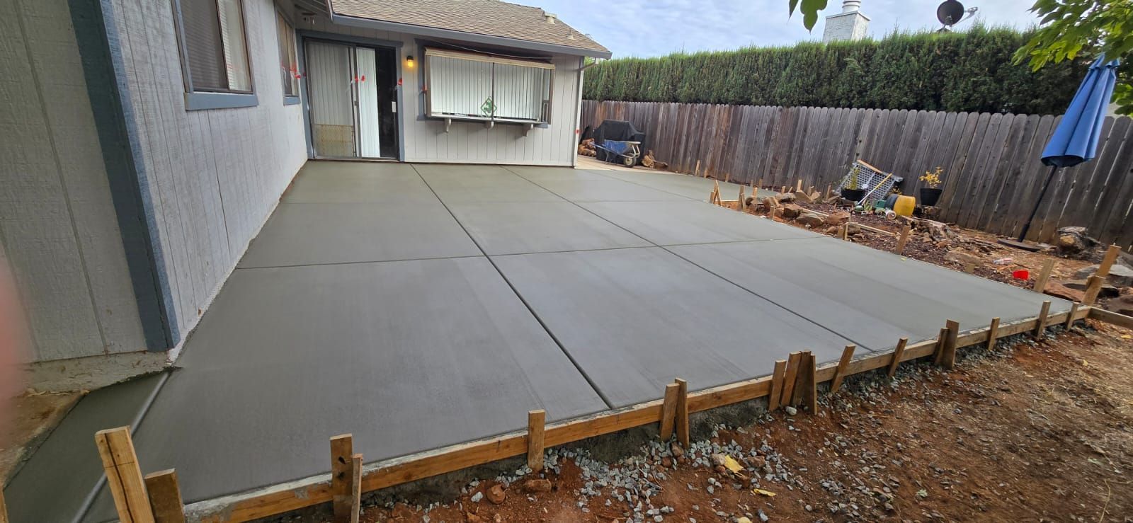 A newly poured concrete patio in a backyard with wooden formwork along the edge and a fence in the background.