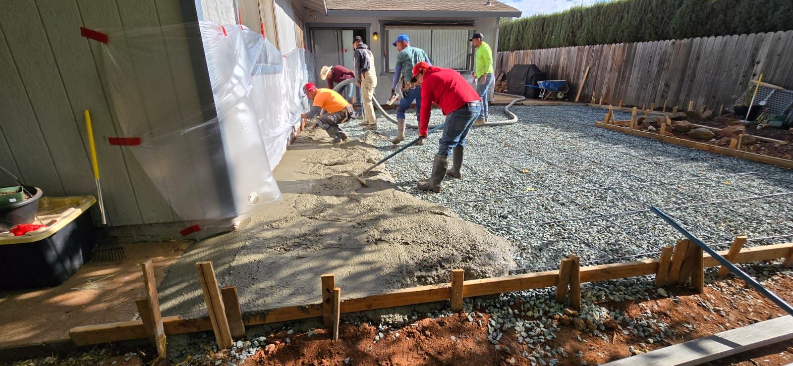 Construction crew in work clothes smoothing wet concrete in a gravel yard near a house.