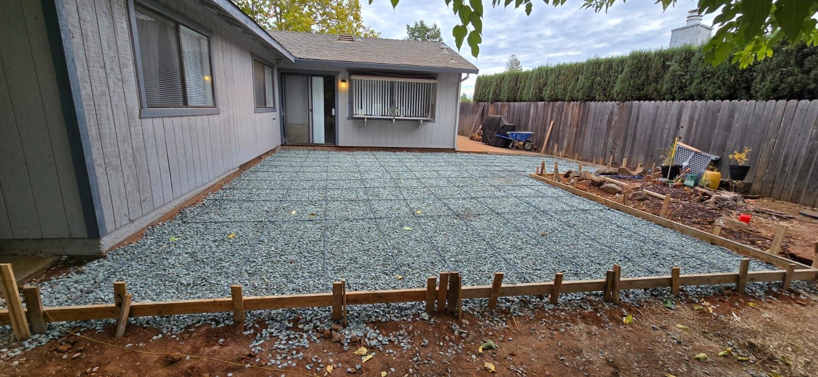 A backyard construction site featuring a gravel-filled patio area defined by wooden formwork for a new concrete slab.
