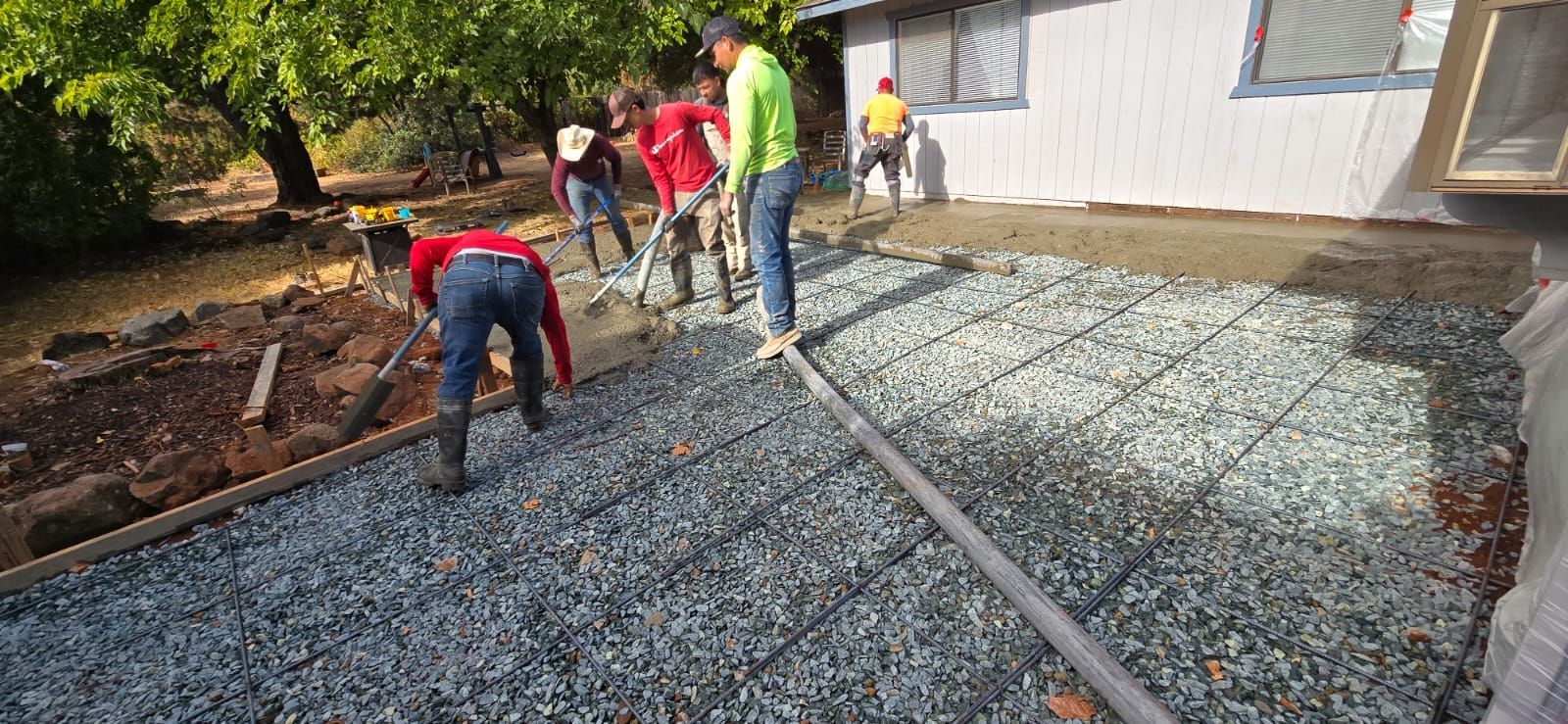 Four workers prep a gravel surface with metal grid reinforcement outside a house.