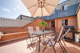 A brick terrace featuring a metal table, four chairs, a beige umbrella, and a barbecue grill under a clear blue sky.