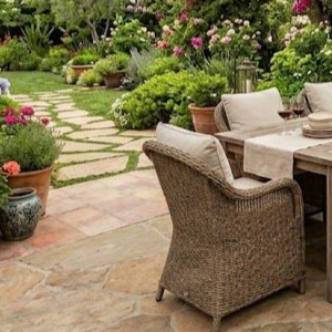A wicker chair with cream cushions sits by an outdoor wooden dining table on a stone patio overlooking a lush garden.