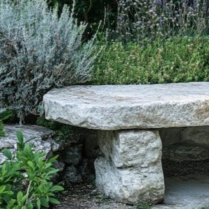 A rustic stone bench sits in a lush garden surrounded by green foliage and lavender.
