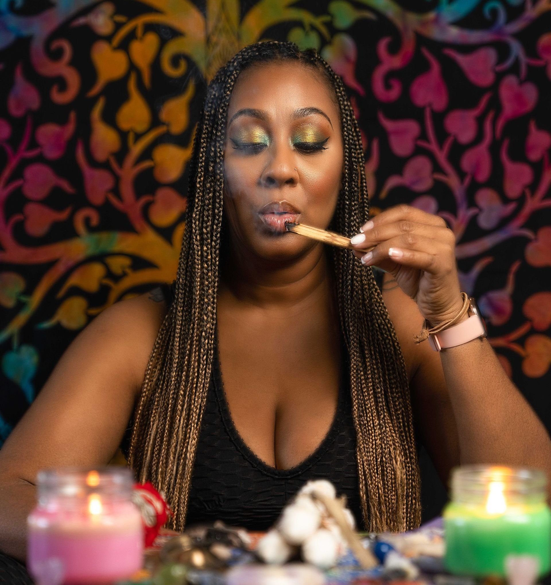 Woman with long braids smoking a stick with colorful eye makeup. Background is a tie-dye design; candles in front.