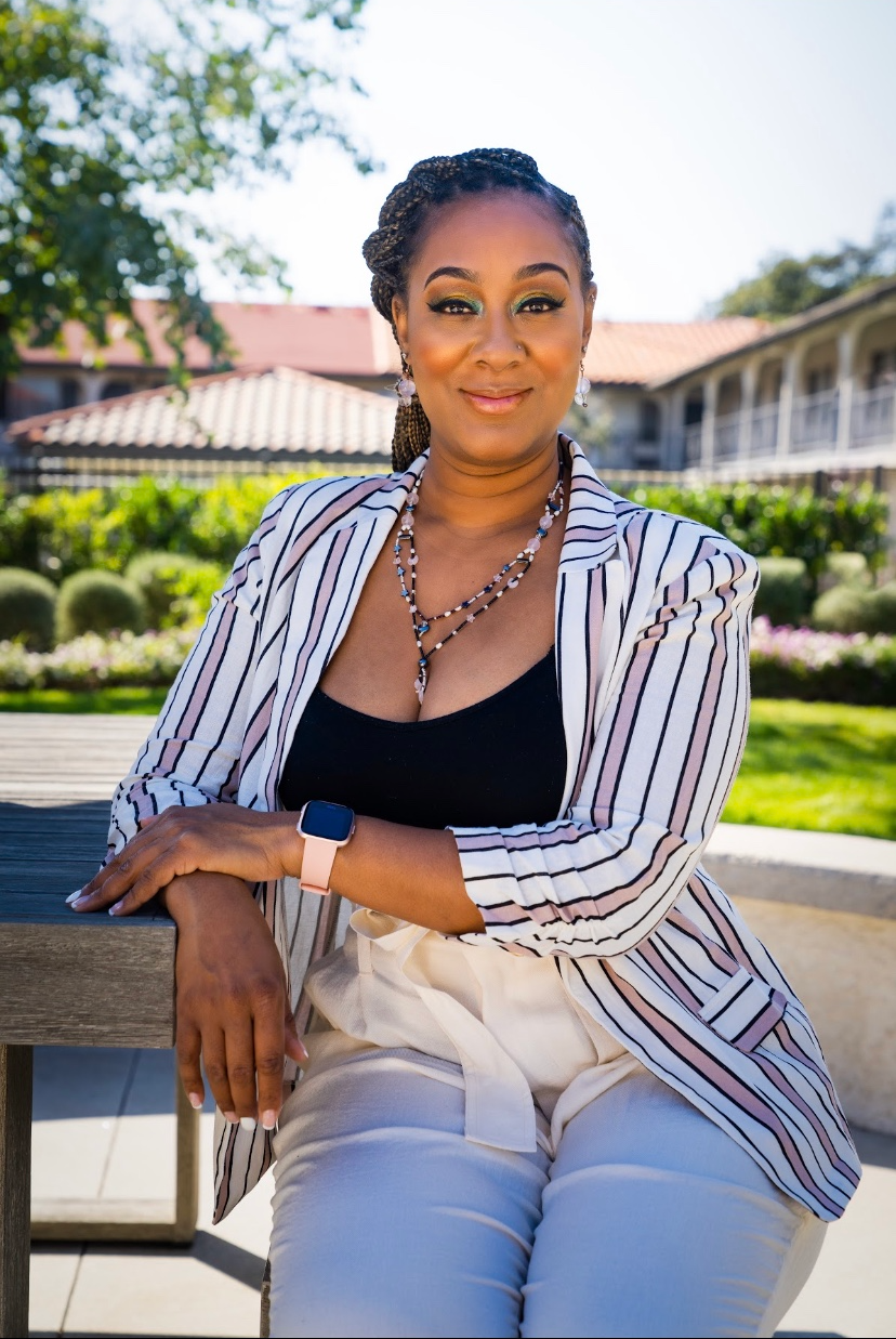 Woman in blazer leans on a table outdoors, smiling. Building and greenery in background.