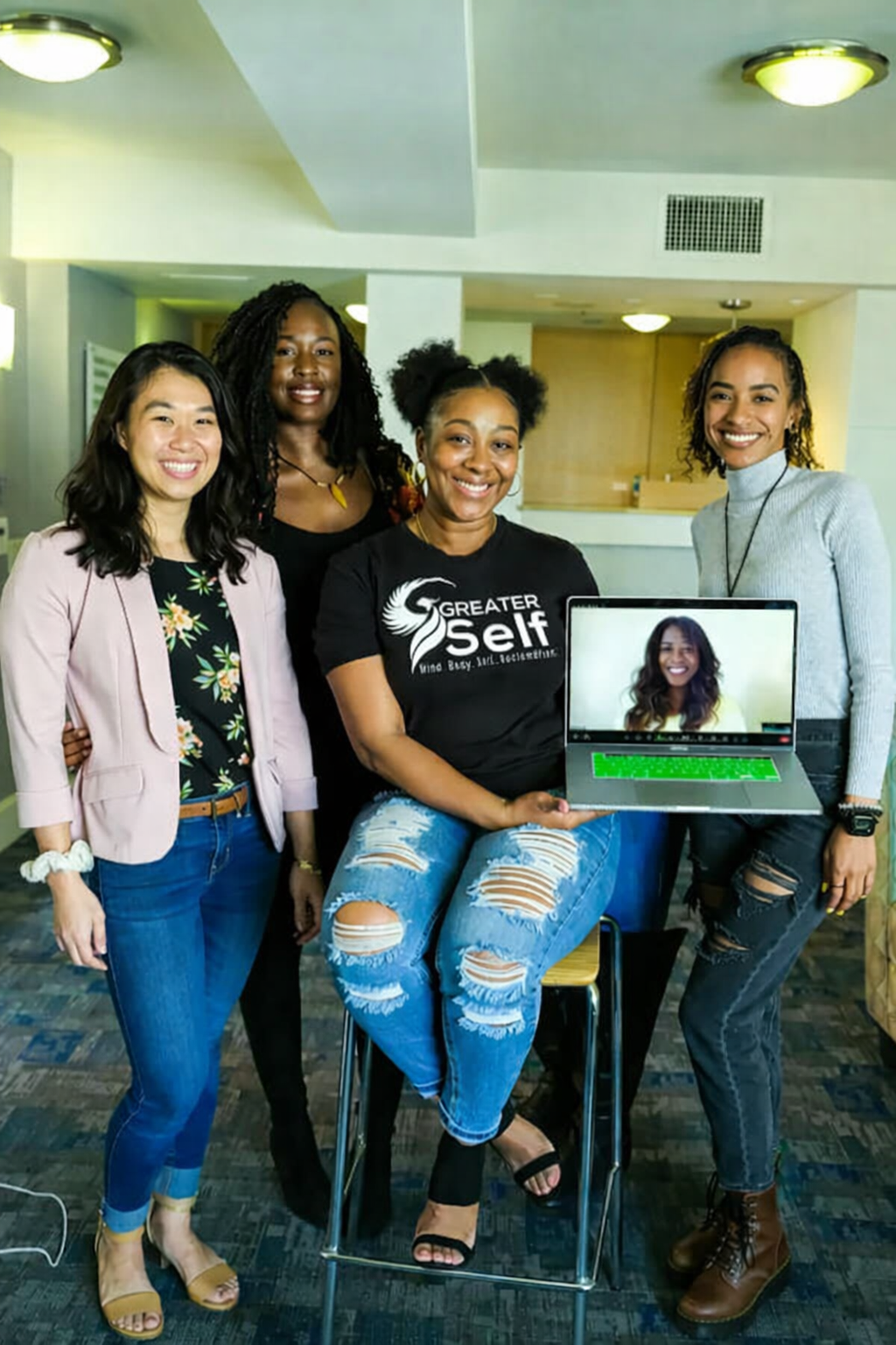 Four people stand indoors, one holding a laptop displaying a woman on screen. Brightly lit room.