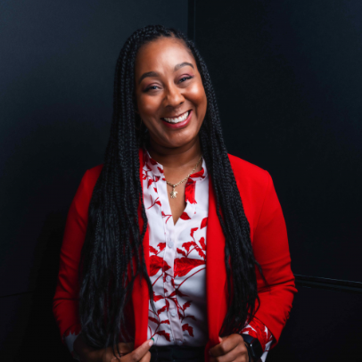 Woman smiling, wearing a red blazer over a white floral print shirt, black braids, against a dark background.