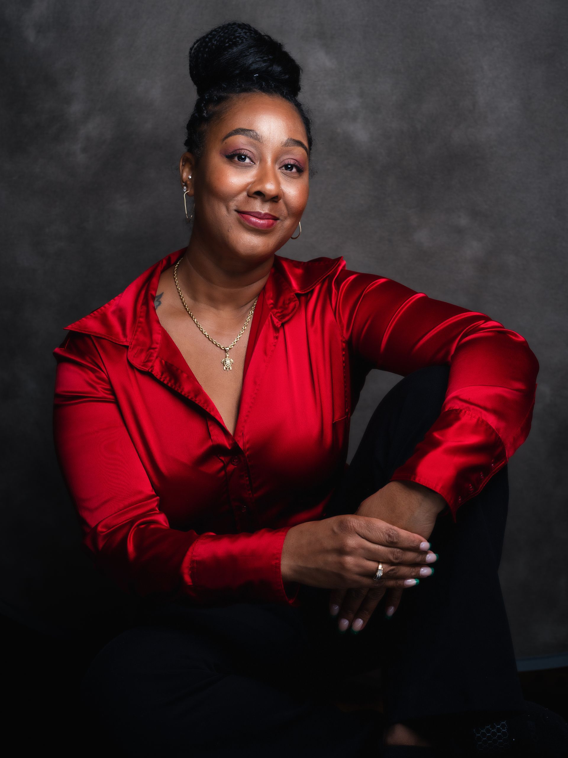 Woman in red satin shirt, black pants, and bun smiles, posing against a gray backdrop.