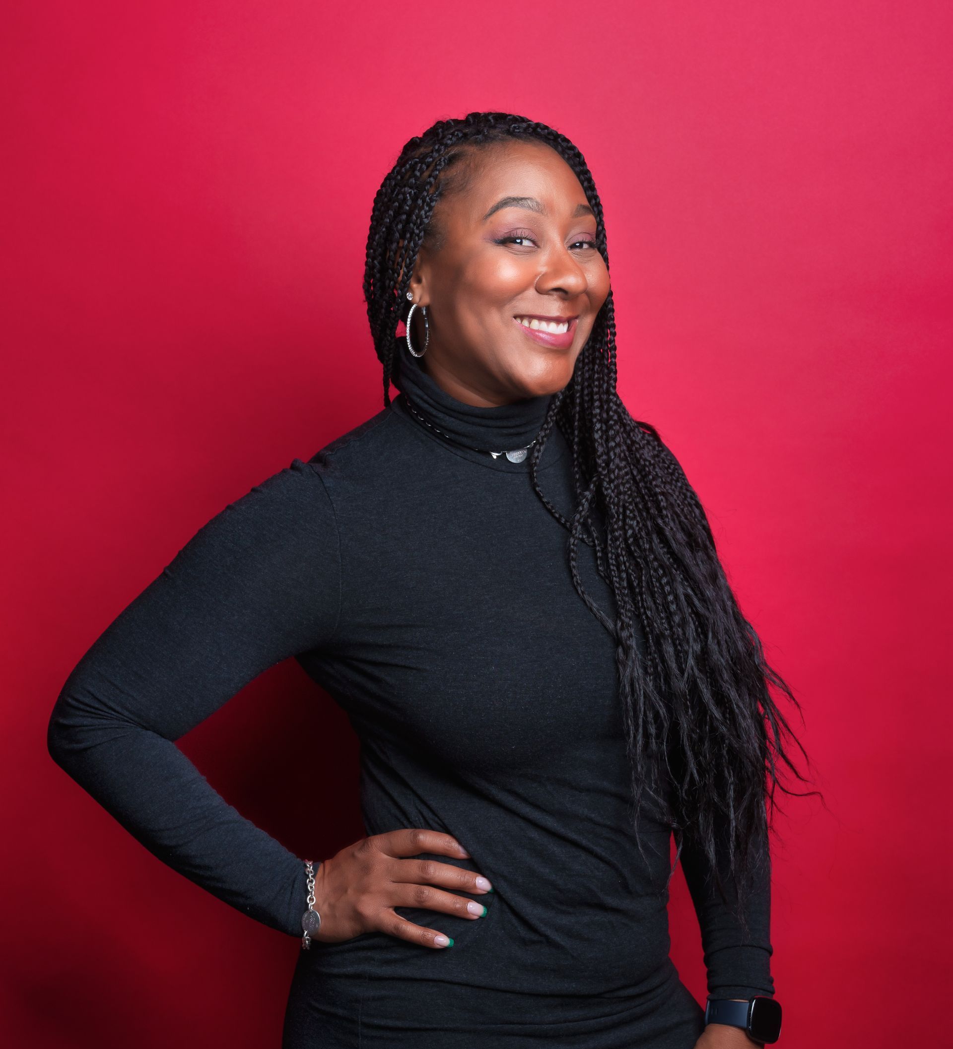 Woman with long braids smiles, wearing a black turtleneck, and poses with a hand on her hip in front of a red background.