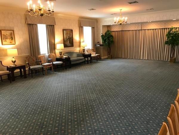 A large, empty event room with beige walls, patterned blue carpet, cream curtains, two chandeliers, and scattered seating.