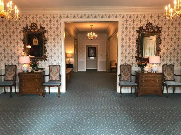 A symmetric room with blue patterned carpet, two mirrors over wooden chests, and four chairs framing a central hallway.