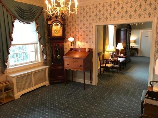 A classic room with a tall grandfather clock, a wooden writing desk, teal carpeting, and a doorway leading to a parlor.