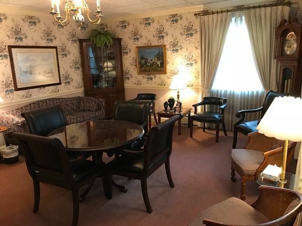 A dining room with a round wooden table, black chairs, floral wallpaper, a grandfather clock, and a window with curtains.