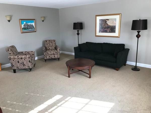 A living room with a dark sofa, two patterned armchairs, a round wooden coffee table, two floor lamps, and two wall photos.