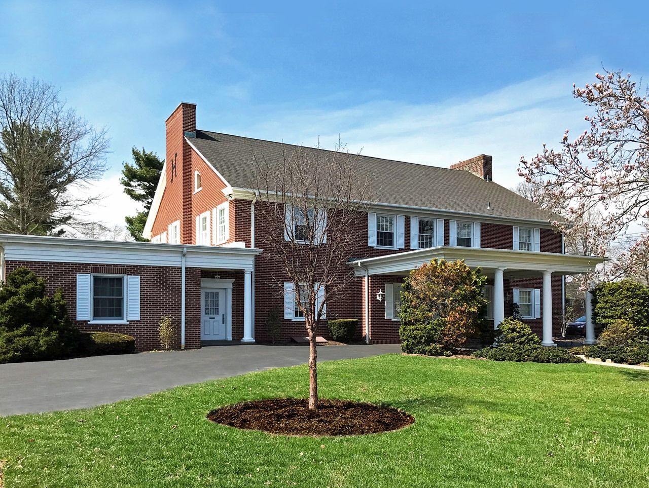 A two-story red brick house with white trim, a covered porch, a green lawn, and a small tree in the foreground.