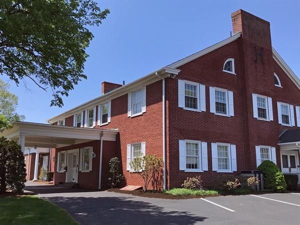 A two-story red brick building with white shutters, a gabled roof, and a covered carport entrance in a parking lot.