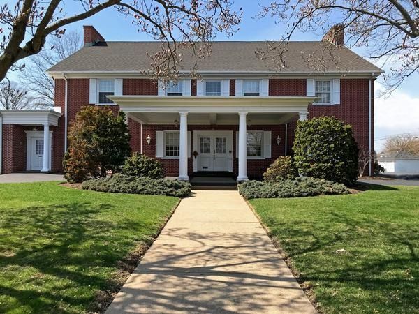A two-story red brick house with a white front porch, a central walkway, and manicured landscaping under a clear sky.