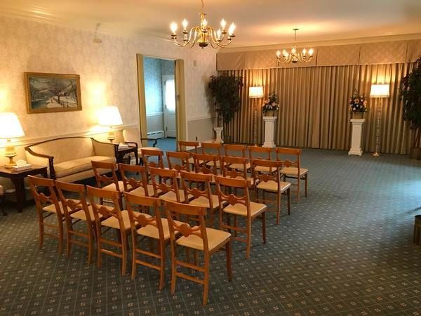 An interior view of a room with rows of wooden chairs, a sofa, chandeliers, floor lamps, and tan curtains.