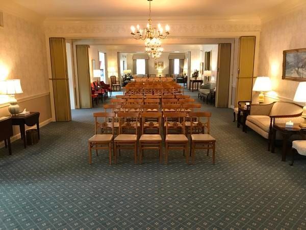 A formal room arranged with rows of wooden chairs, flanked by lamps, sofas, and a blue-patterned carpet.