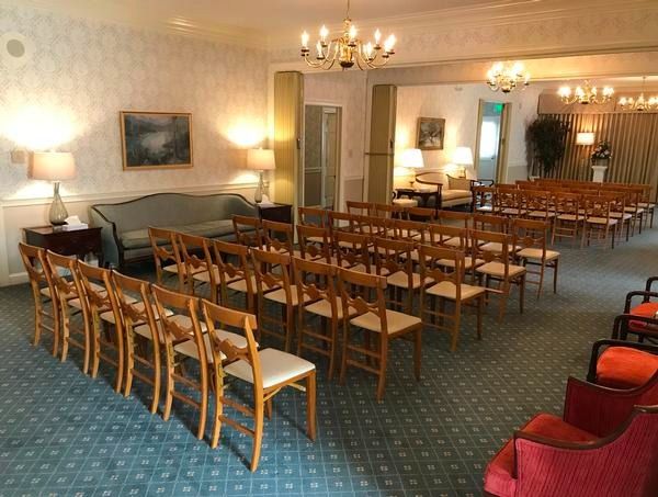 A formal event space with rows of wooden chairs, a light blue carpet, and a large mirror reflecting a seating area.