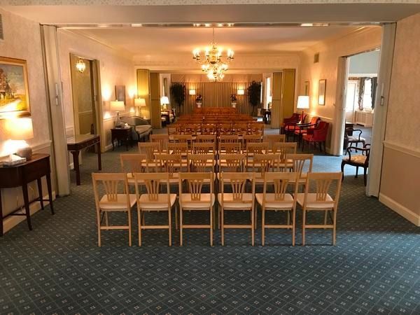 An interior view of a room set up for an event, featuring rows of light-wood chairs on a blue patterned carpet.