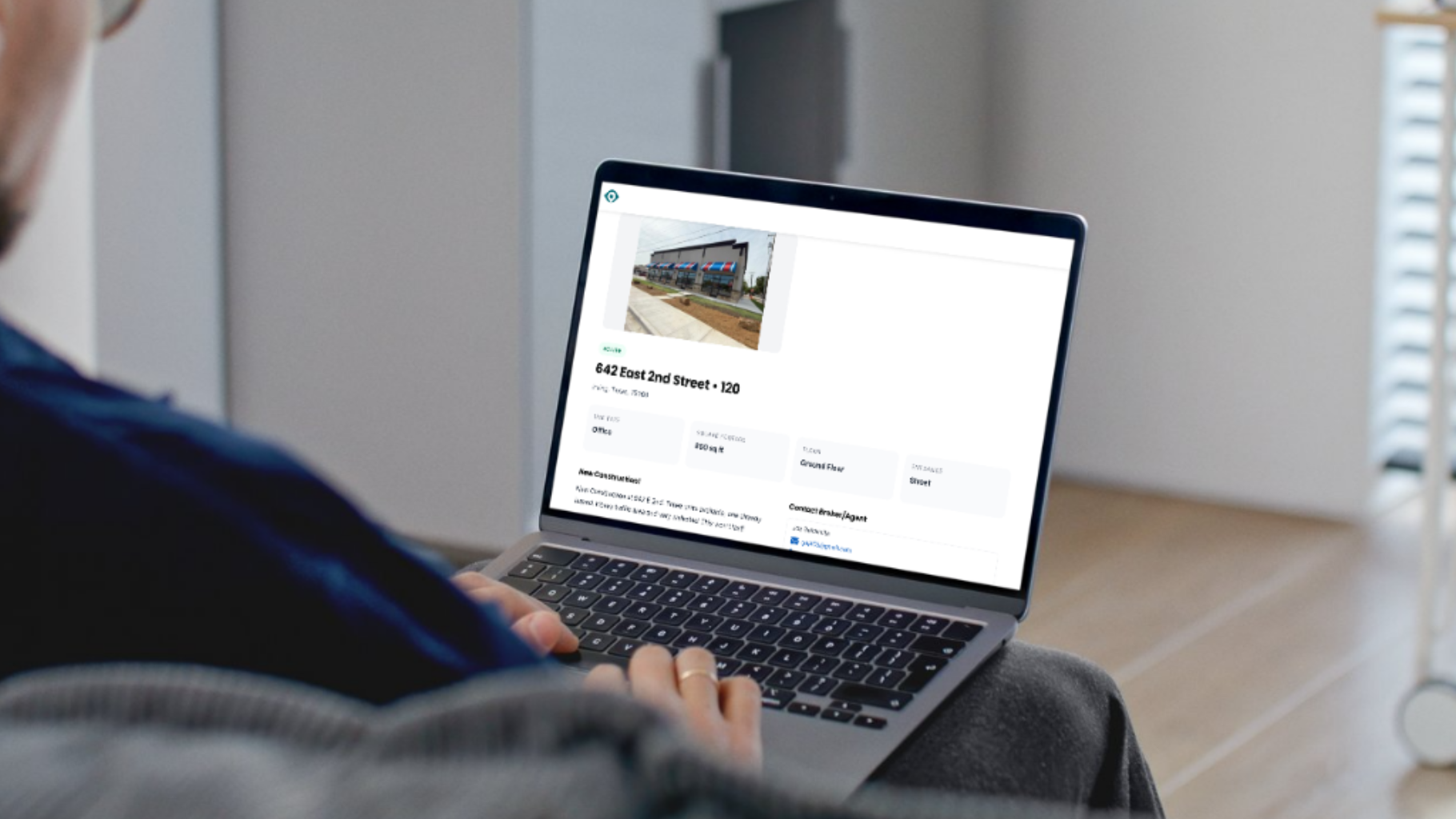 A person uses a laptop displaying a real estate listing for a building, seen from an over-the-shoulder perspective.