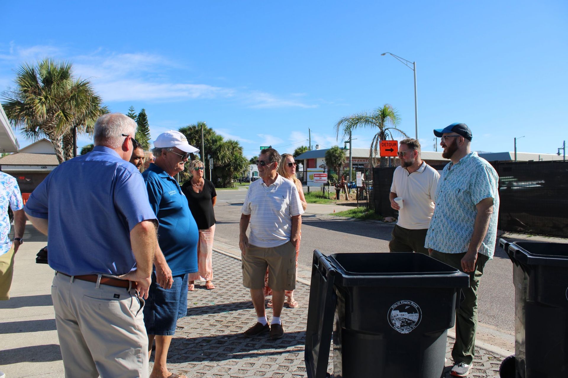 A group of people standing on a street next to large trash bins during a sunny day.