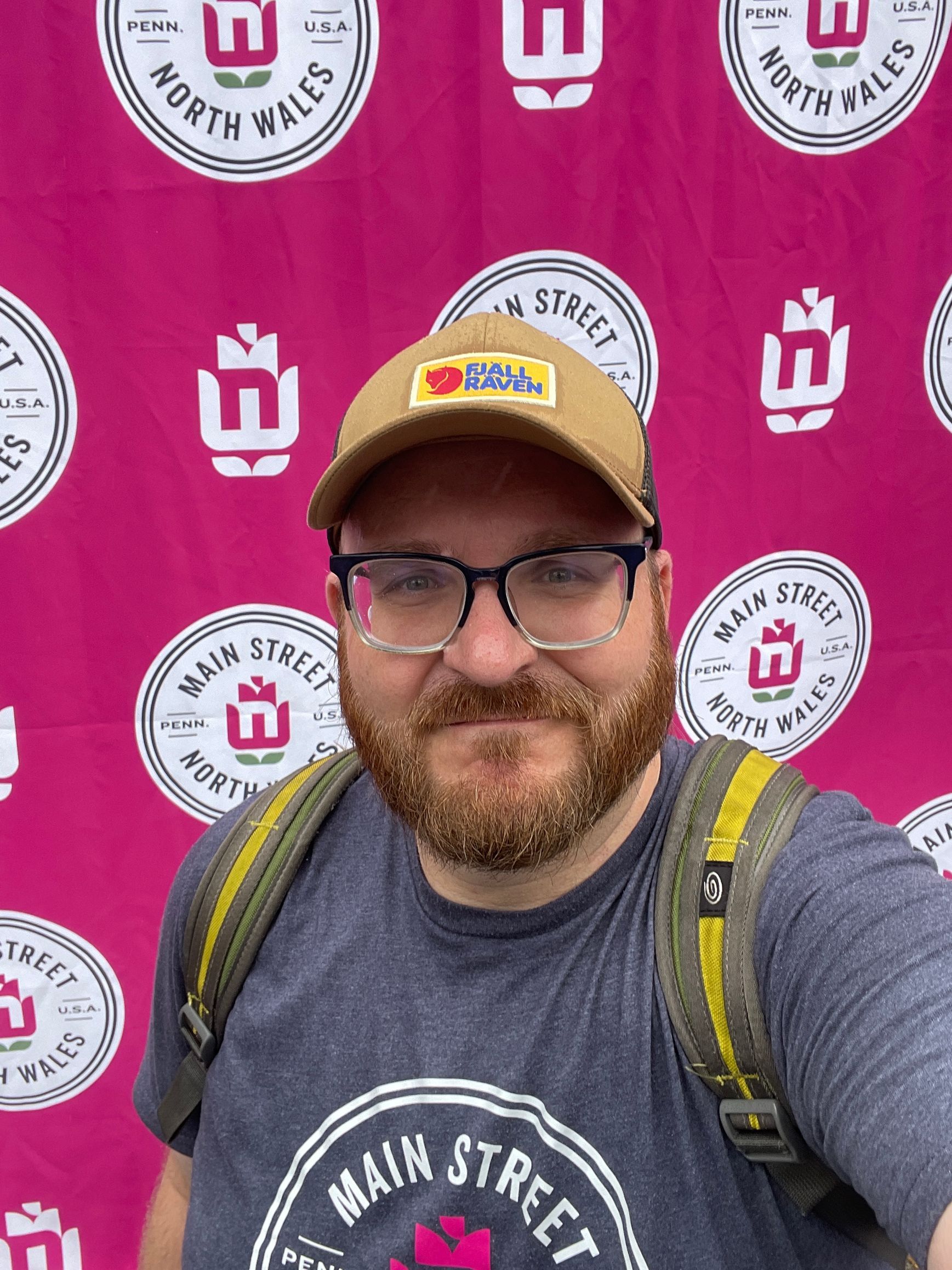 A person with a beard and glasses wears a baseball cap and backpack in front of a pink Main Street North Wales backdrop.